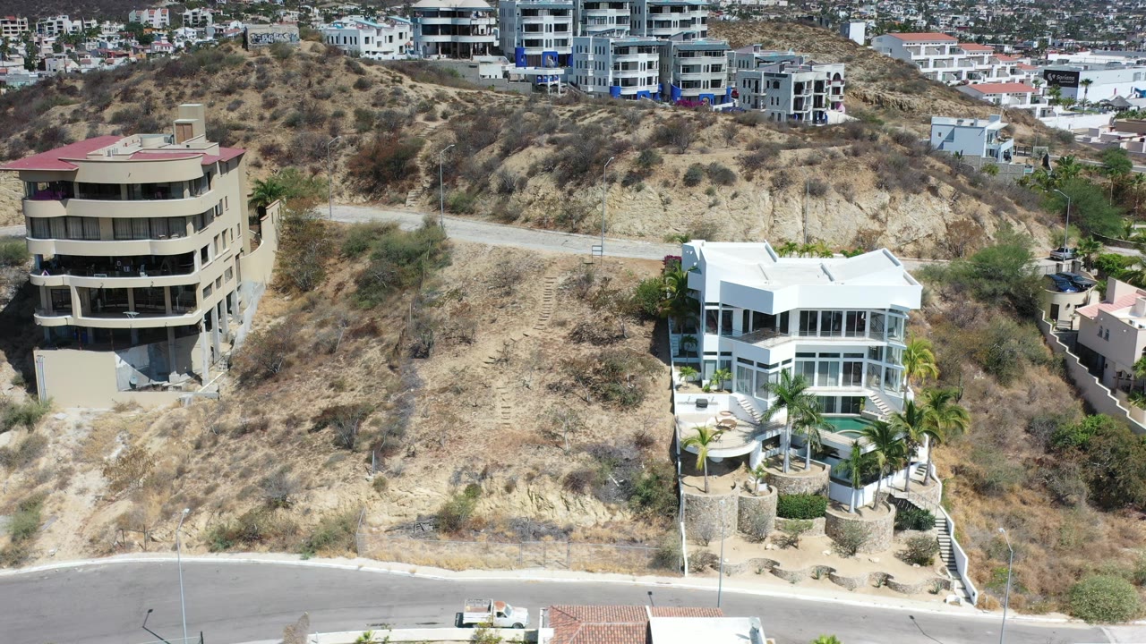 A Los Cabos aerial frame centers dry hillside brush between a tan apartment building and a white glass-fronted home, with city buildings behind and no visible people.