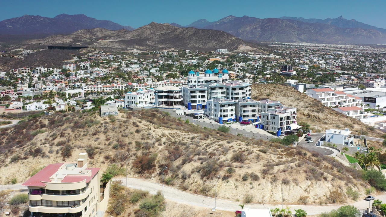 Opening elevated view in Los Cabos, Mexico, with dry scrub hills in front, a white resort complex below, town buildings across the valley, distant mountains, and no visible people.