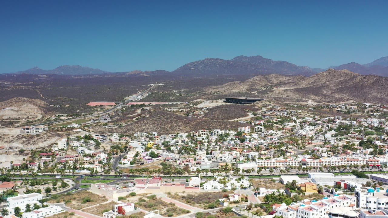 Early aerial view of Los Cabos, Mexico, showing a dense band of white buildings below scrub-covered hills and a clear blue sky, with no visible people.