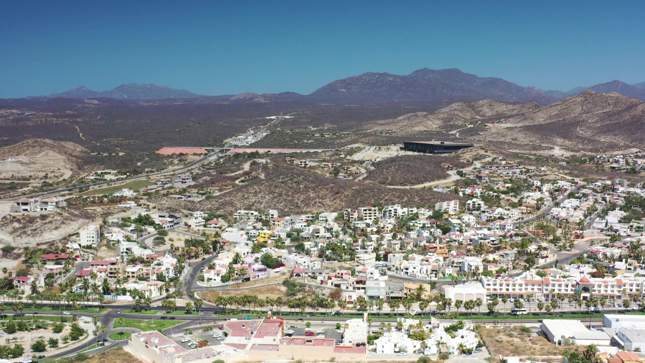Second aerial frame in Los Cabos showing a broad sweep of light-colored houses, a main road, small cars, dry ridges, distant mountains, and no visible people.