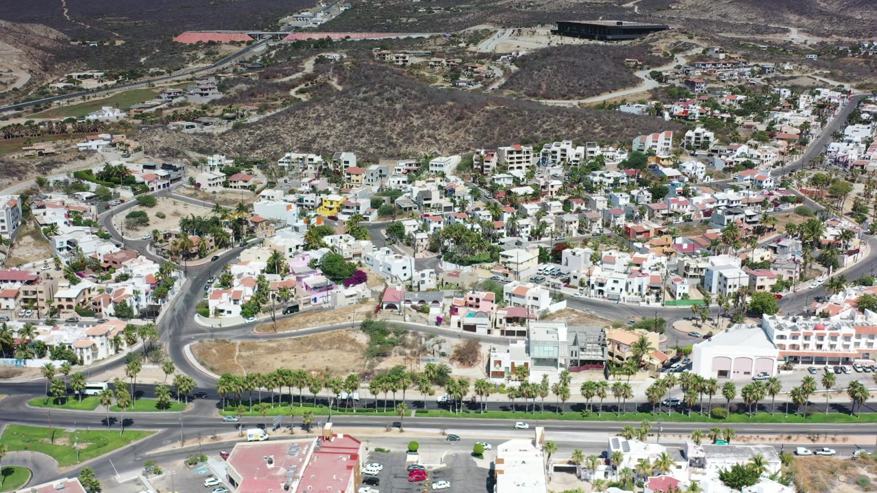 High aerial frame over Los Cabos, Mexico showing a multi-lane road with cars, rows of palms, white resort buildings, hillside houses, dry terrain, and no visible people.