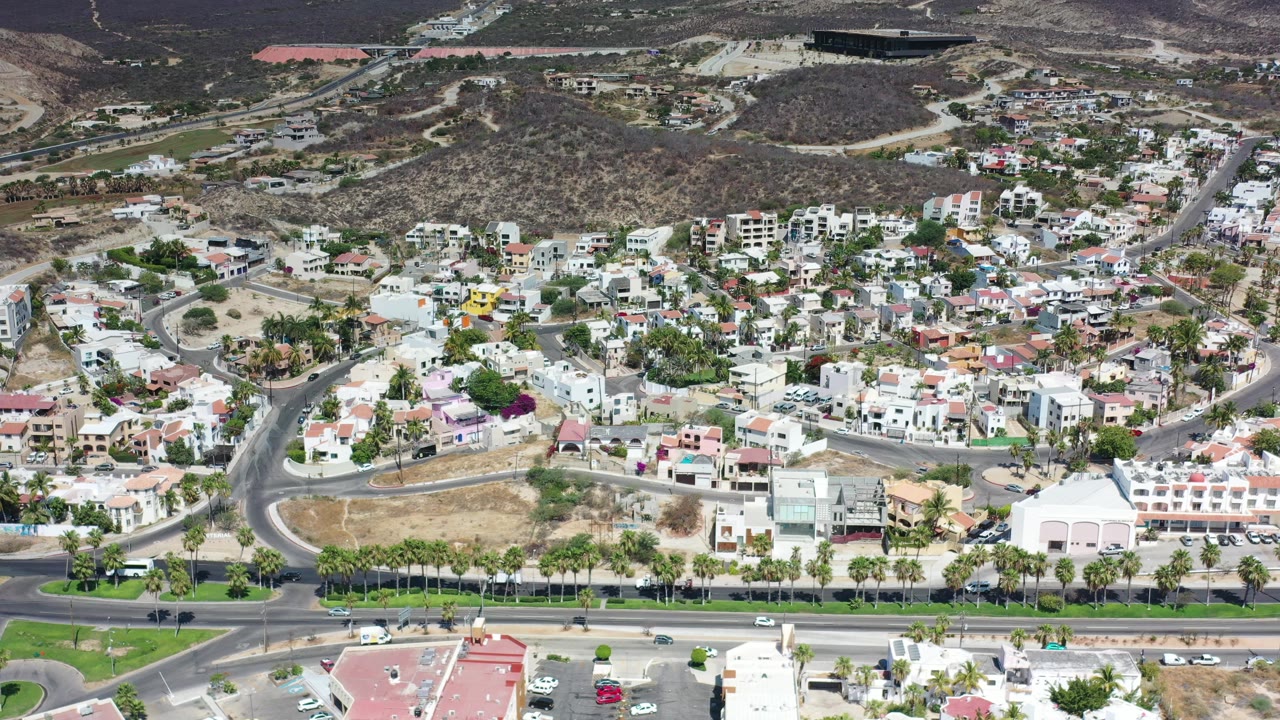 Aerial Los Cabos view one second into the clip, with cars along the road, a green palm-lined strip, white resort structures, clustered hillside homes, brown hills, and no visible people.