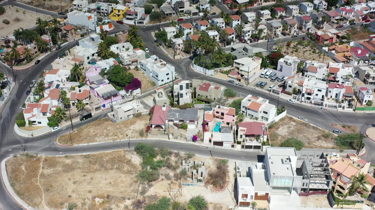 This Los Cabos, Mexico frame looks down on intersecting roads, compact homes, parked vehicles, palms, and dry tan parcels with no visible people.