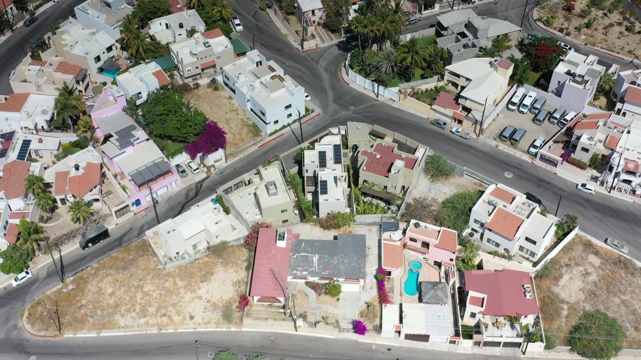 Overhead frame in Los Cabos, Mexico showing a road intersection, white residential buildings, parked cars along curbs, palms, dry ground, and no visible people.