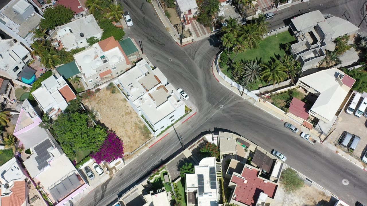 Overhead view of a Los Cabos residential intersection with white flat-roof homes, parked cars, palm trees, a small blue pool, and no visible people.