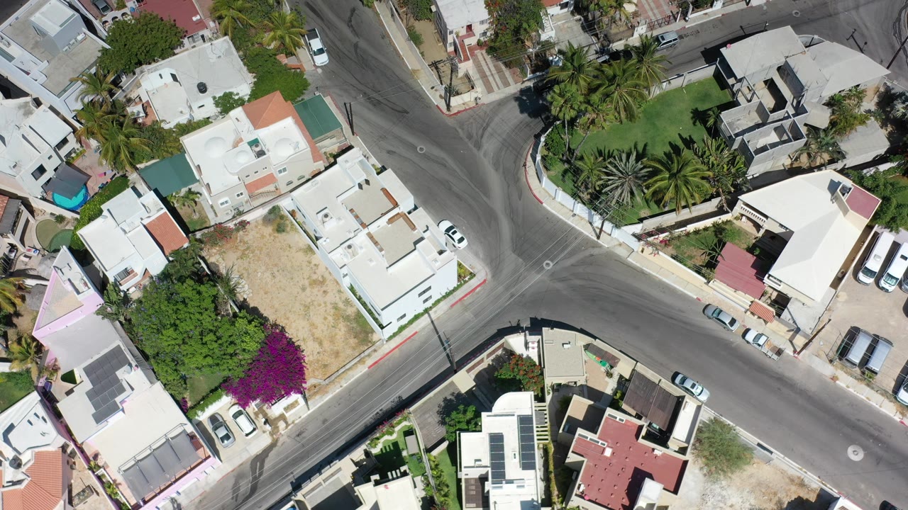 Top-down Los Cabos neighborhood frame centered on a broad road junction, pale rooftops, garden walls, parked vehicles, palms, and no visible people.
