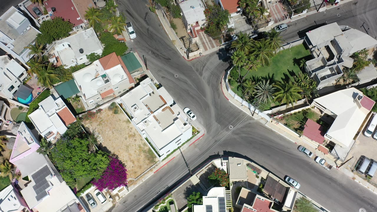Aerial frame of residential streets, flat roofs, palms, and parked cars in bright sun.