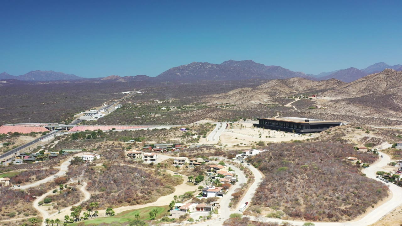 Wide aerial frame over Los Cabos showing dry hills, pale roads, scattered homes, a large dark building, distant mountains, and no visible people.