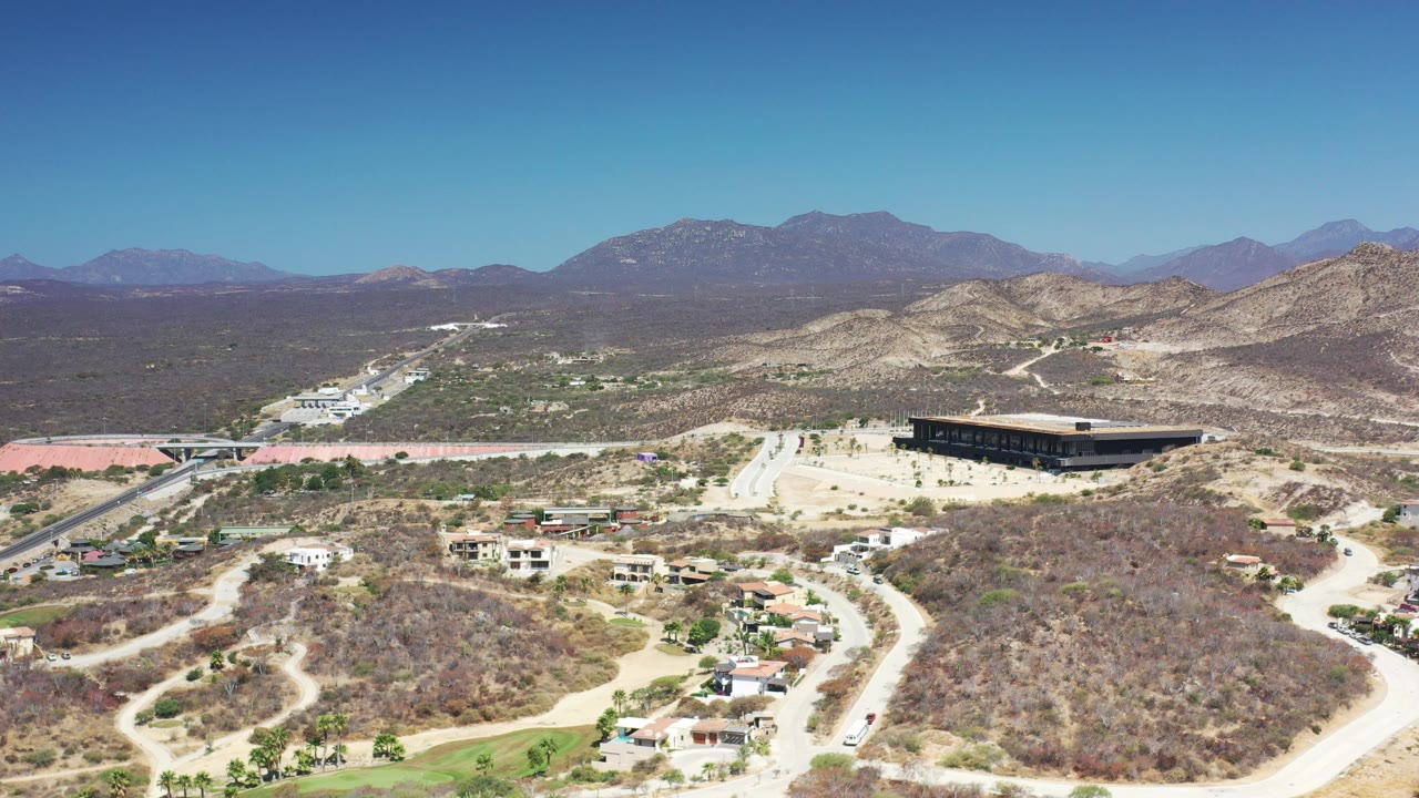 Aerial view in Los Cabos with a paved road crossing the scene, hillside homes below, a dark rectangular building to the right, and no visible people.