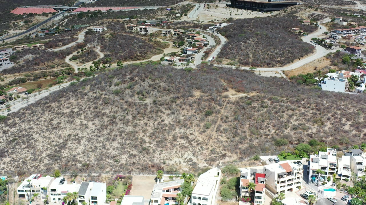 Los Cabos, Mexico aerial view showing a broad scrub-covered hill, curving dirt roads, a few homes on the upper slopes, and apartment-style buildings entering the foreground.