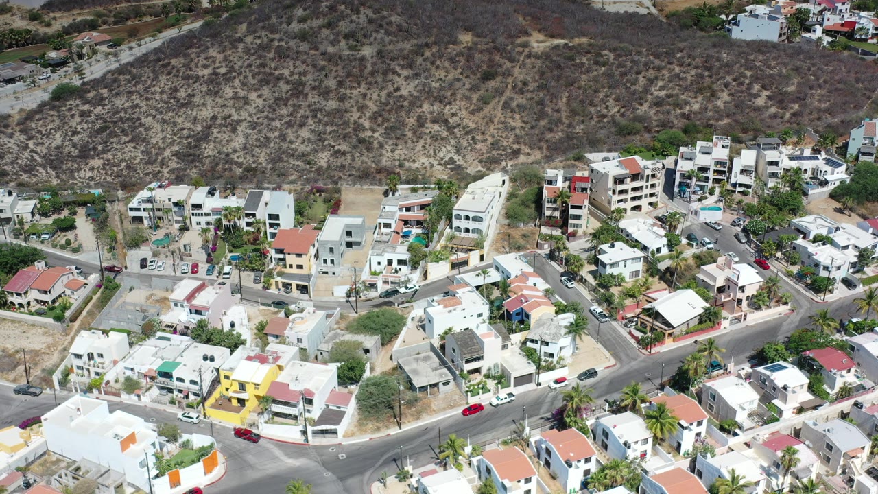 Aerial frame of Los Cabos residential blocks with white rooftops, red tile roofs, a yellow house, paved streets, sparse traffic, palm trees, and dry scrubland rising behind, with no visible people identifiable in the frame.