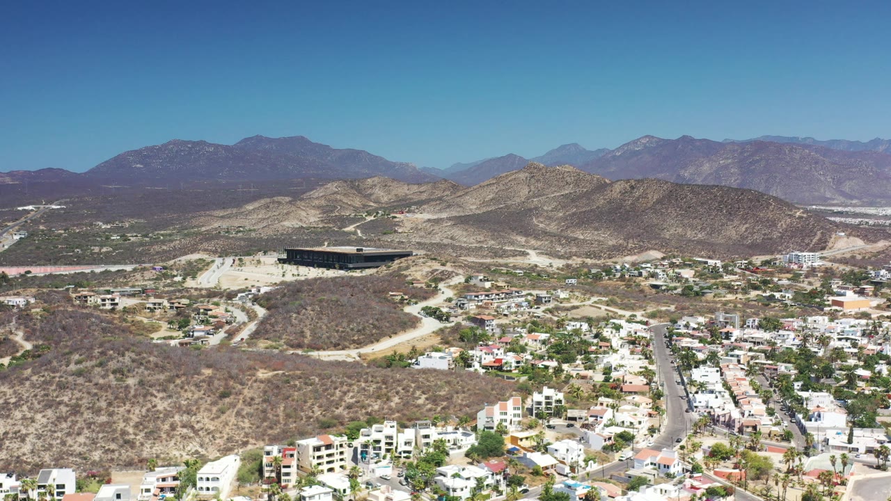 Wide aerial frame in Los Cabos, Mexico, showing pale homes and streets in the foreground, scrub-covered hills across the middle distance, a dark rectangular building, mountains, and no visible people.