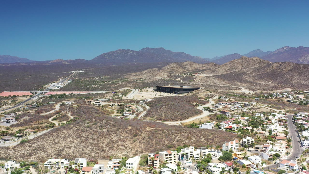 Opening aerial frame over Los Cabos, Mexico, with white homes at the bottom edge, scrub-covered hills, pale roads, a dark rectangular building, distant mountains, and no visible people.