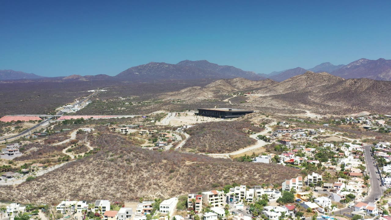 Second frame of the Los Cabos aerial shows more hillside road detail through the center, residential blocks along the lower edge, layered mountains in back, and no visible people.