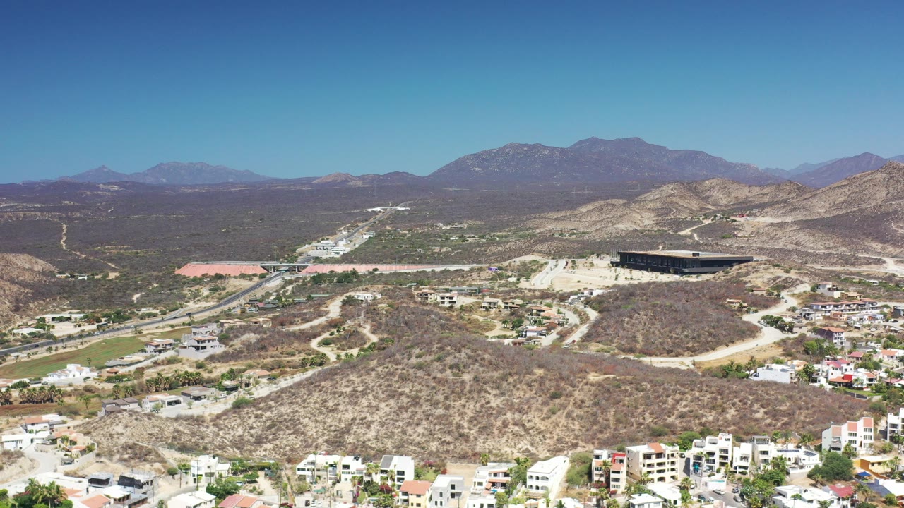 Aerial view of dry hills, road, homes, and distant mountains under a blue sky.