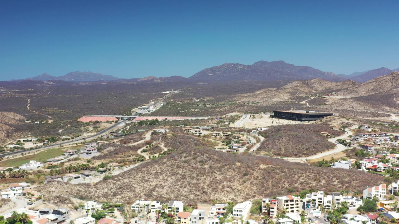 Los Cabos aerial frame showing pale desert slopes, low vegetation, a road corridor, white residential buildings, a dark structure on the hillside, and no visible people.