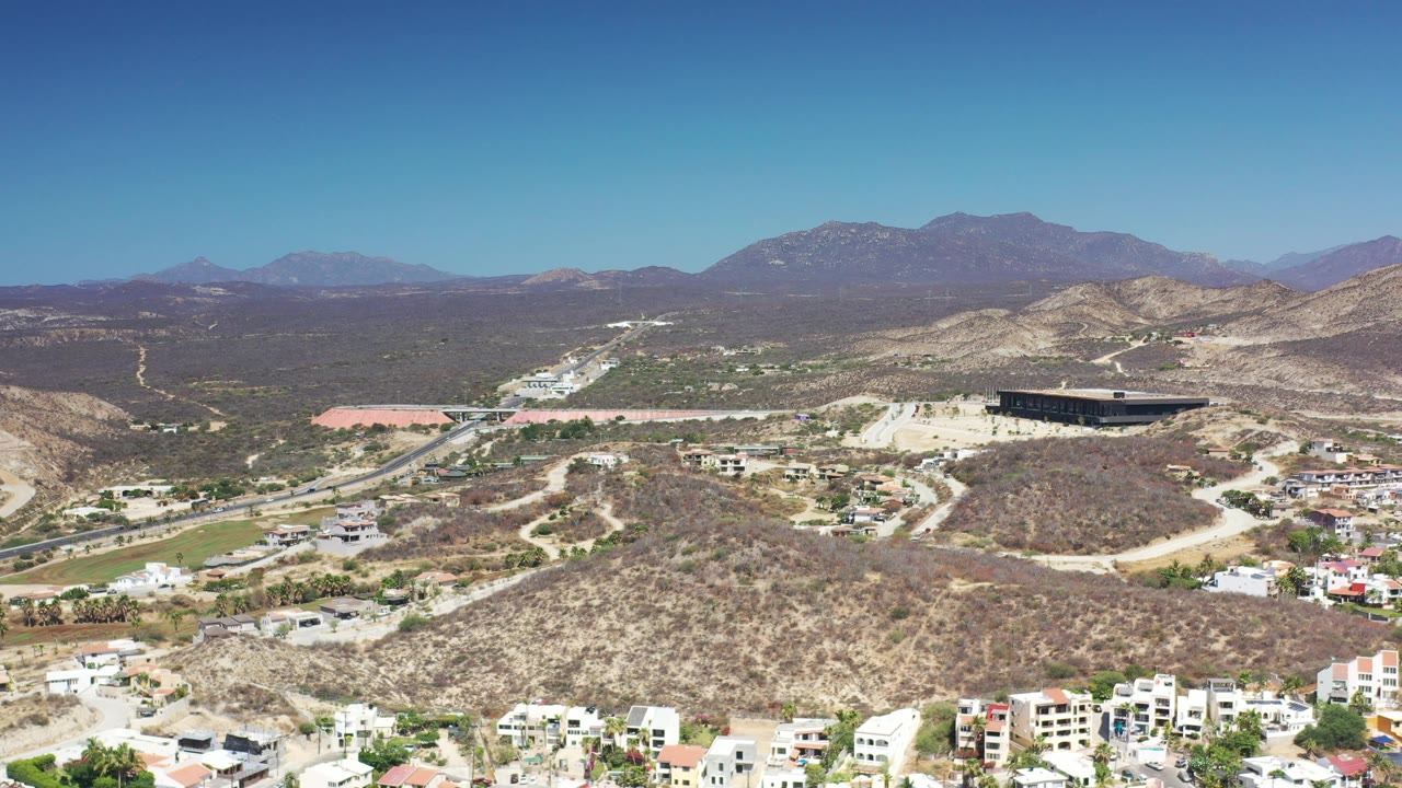 Wide Los Cabos aerial view with dry brown hills in the foreground, a road network through the valley, scattered buildings, mountain ridges, and no visible people.