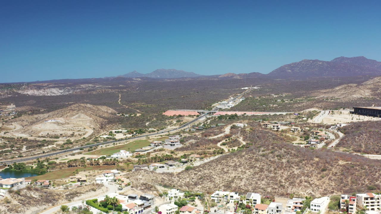 Aerial frame over Los Cabos desert hills, showing a central roadway, pale construction areas, small residential buildings, mountain ridges, and no visible people.