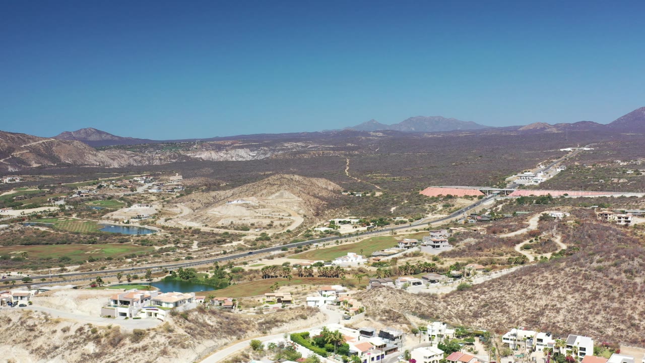 The second frame keeps a high Los Cabos aerial composition with scrub-covered slopes, light-colored homes, curving roads, green fairway-like landscaping, small water areas, and no visible people.