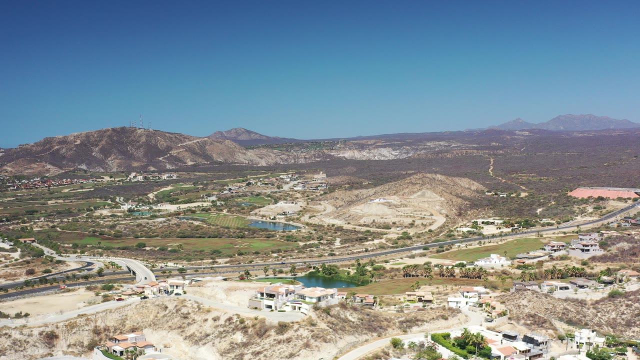 Wide aerial view of dry hills, roads, fields, and distant mountains under a blue sky.