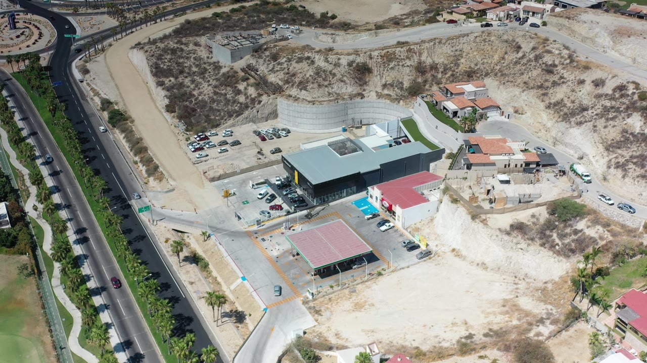 Early aerial frame in Los Cabos, Mexico, with the dark-roofed building centered lower in view, cars parked near it, hillside homes behind, and no visible people.