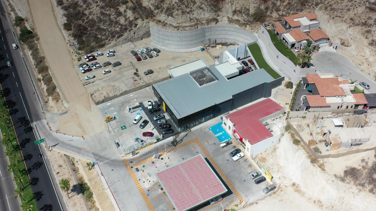 Los Cabos aerial frame showing a pale-roofed modern building centered between roadside parking, a red-roofed structure, nearby homes, dusty lots, and no visible people.