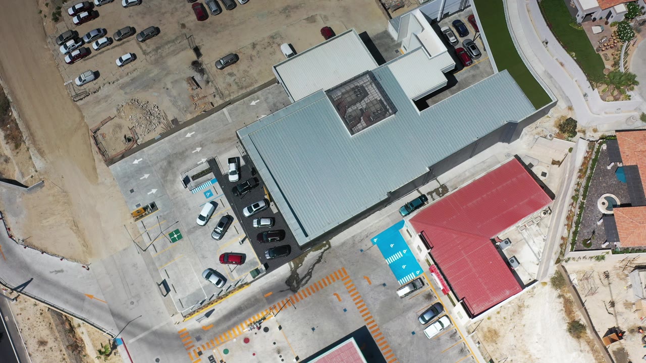 Vertical aerial frame over an industrial park in Los Cabos, Mexico, showing a large gray roof, adjacent parked cars, a red-roofed building, dry ground, access roads, and no visible people.
