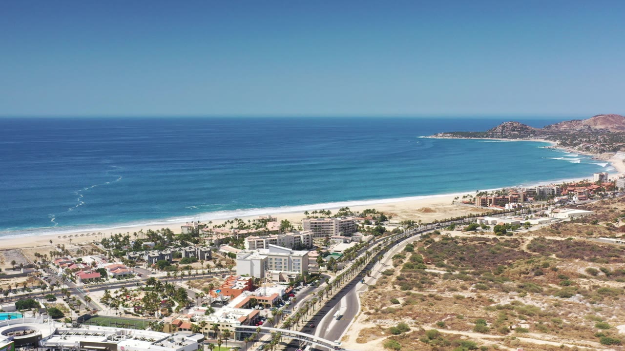 Aerial view of a sandy beach, blue ocean, coastal buildings, roads, and dry hillside.