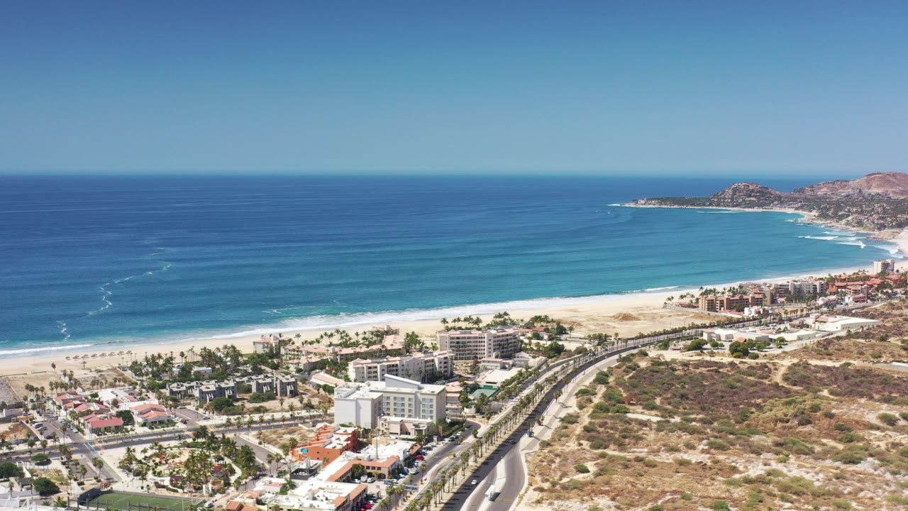 Aerial view over Los Cabos, Mexico with blue ocean, white surf, a sandy beach, beachfront buildings, roads, palms, and dry hillside terrain in bright daylight.