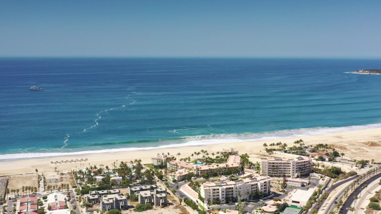 Aerial view of beach buildings, surf, blue sea, boats, and horizon.
