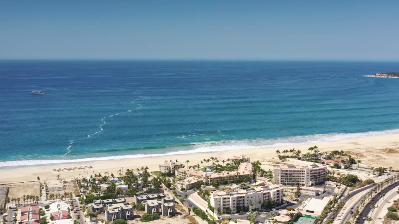 Aerial view in Los Cabos, Mexico, holding on the beachfront district, with a curved road at lower right, resort-like buildings, surf bands, offshore boats, and no visible people.