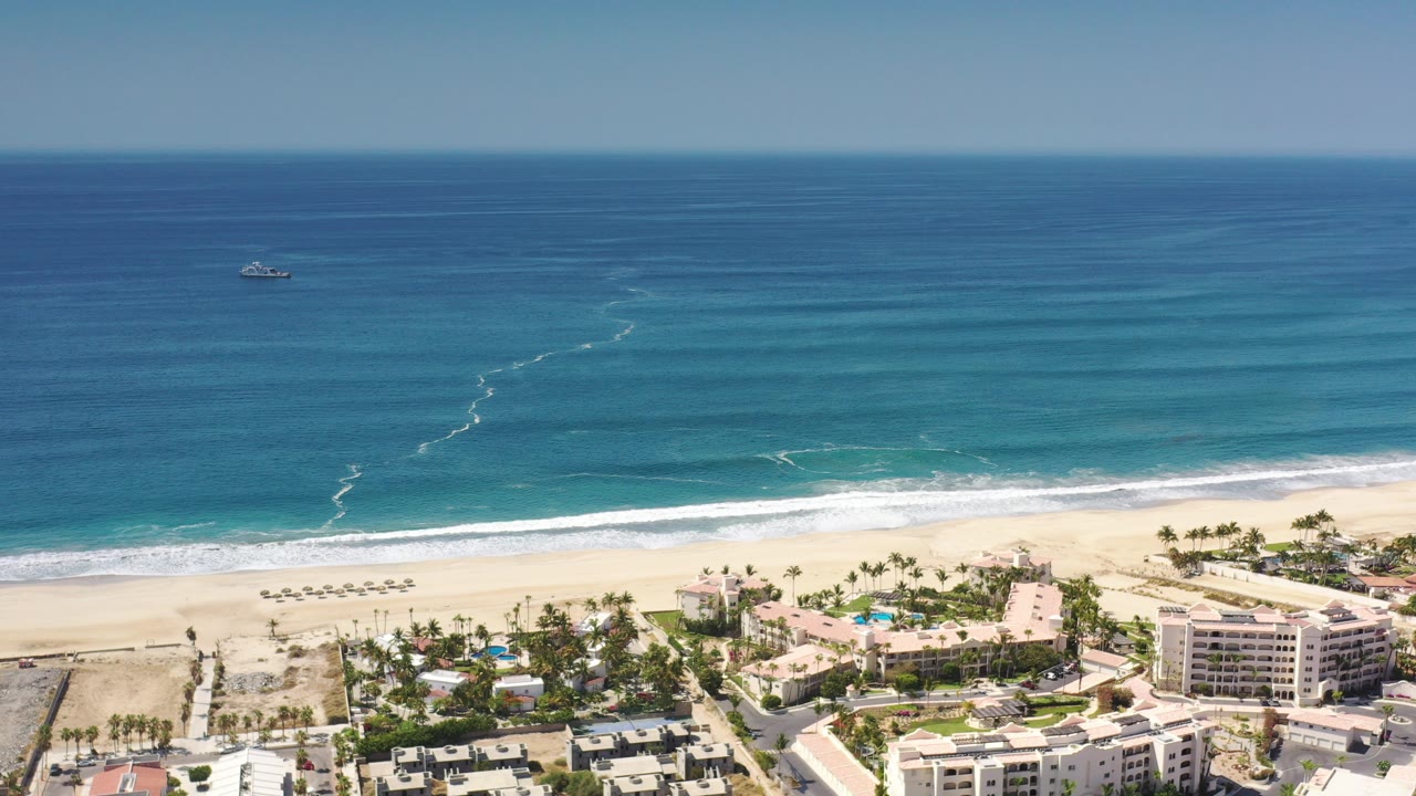 Aerial frame over Los Cabos, Mexico showing deep blue ocean, pale surf, a wide sandy beach, palm-lined resort buildings, pools, and no visible people at the start of the clip.