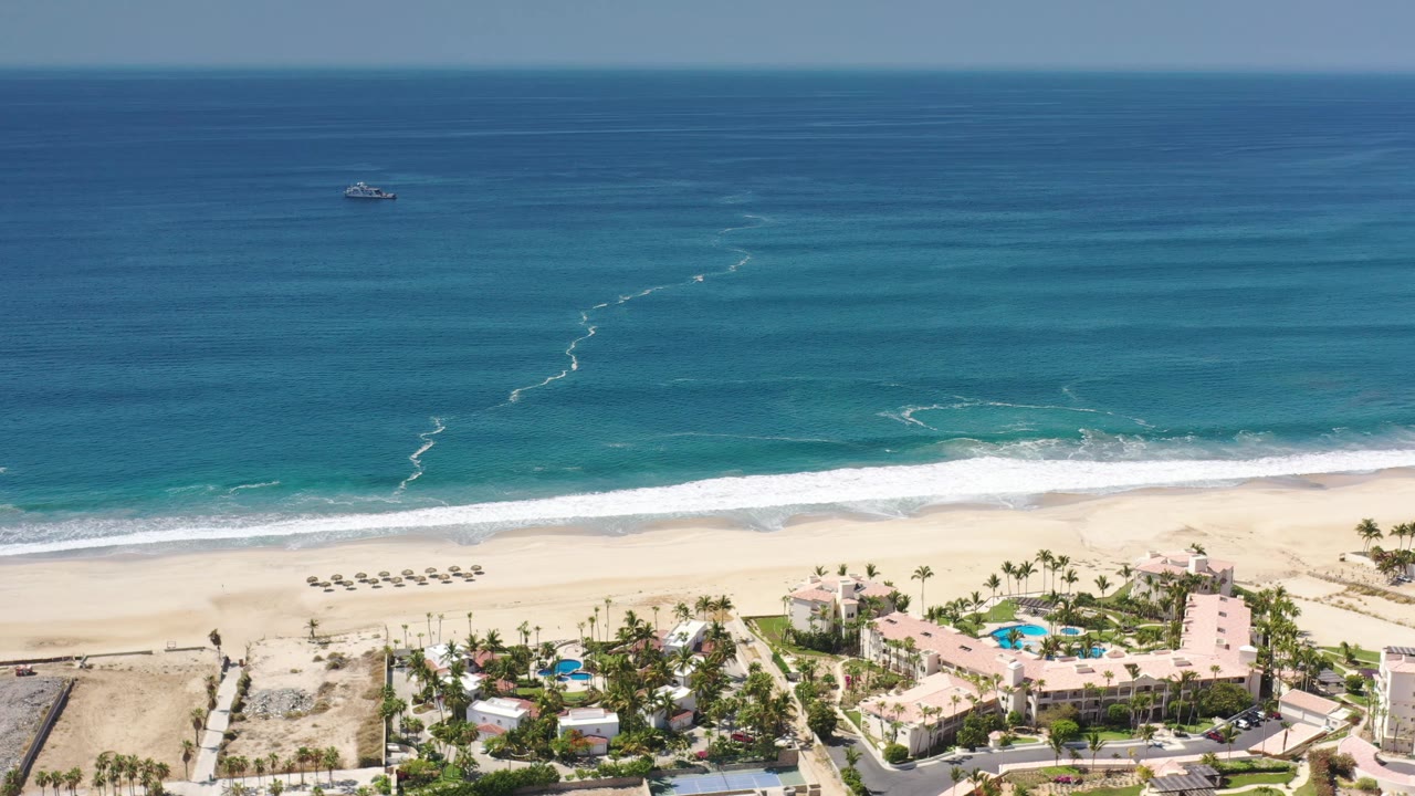 Aerial Los Cabos resort view with the small boat near the upper ocean, white water curling toward the beach, umbrellas on the sand, and no visible people.
