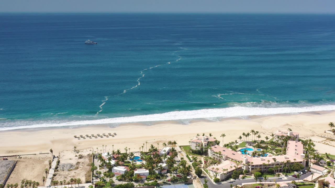 Drone-style view of surf, beach umbrellas, resort pools, and buildings.