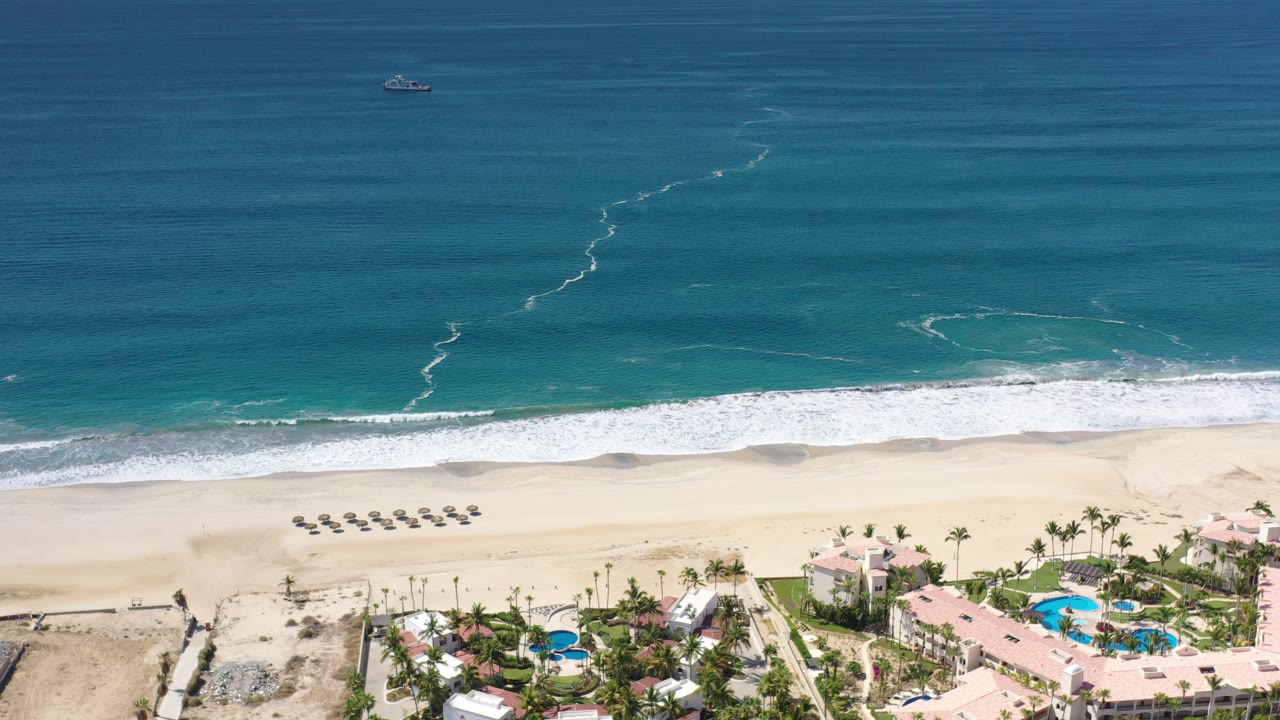 Aerial view of turquoise ocean, white surf, empty beach loungers, and resort pools.