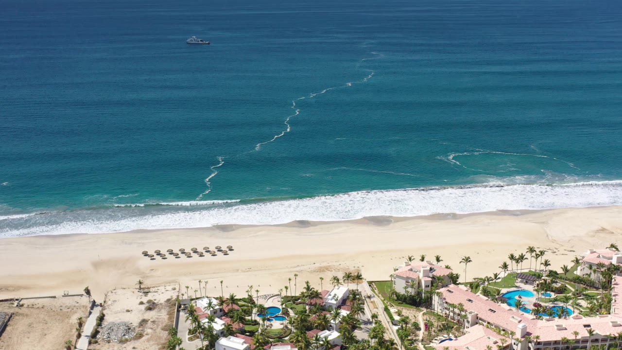 Drone-height view in Los Cabos with darker blue ocean at the top, foamy surf near shore, a wide empty sand strip, aligned loungers, resort roofs, swimming pools, and no visible people.