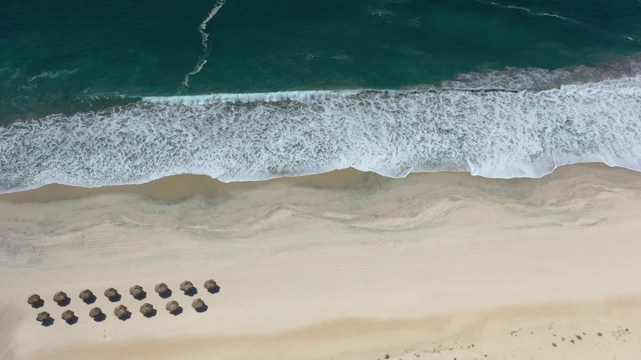 Aerial frame from Los Cabos, Mexico showing a wide band of white foam along the beach and a compact umbrella cluster lower in frame, no visible people.