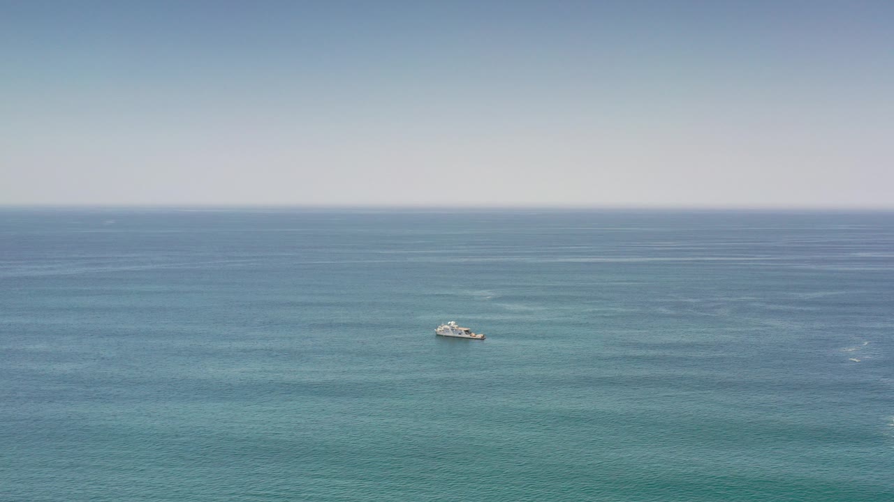 A distant white boat sits left of center on blue ocean near Los Cabos, Mexico, with pale sky above and no visible people.