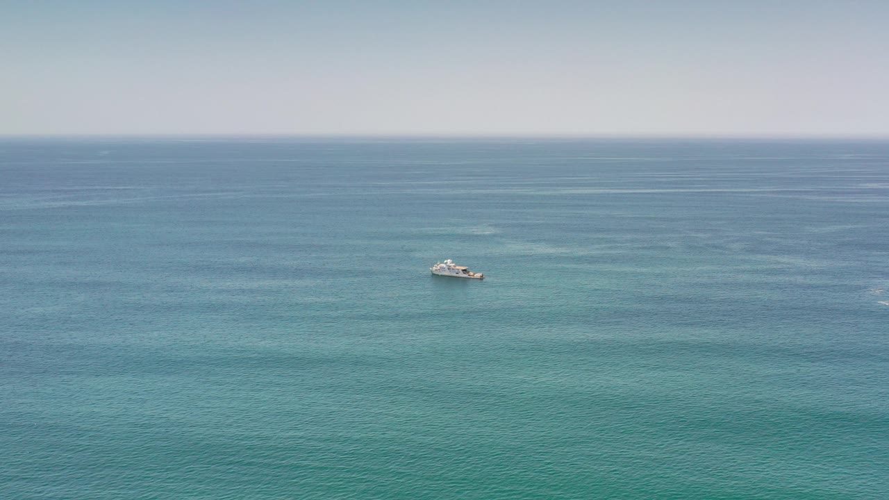 Wide Los Cabos ocean frame with a small white boat left of center, layered blue-green water, a pale sky, and no visible people.