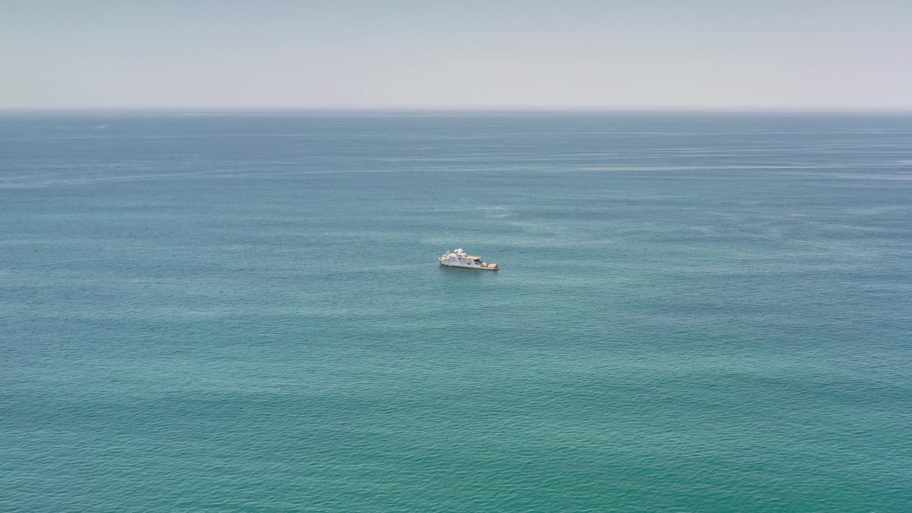 At 0:01, the distant boat remains centered in open Los Cabos water, with subtle turquoise patches below a flat pale sky and no visible people.