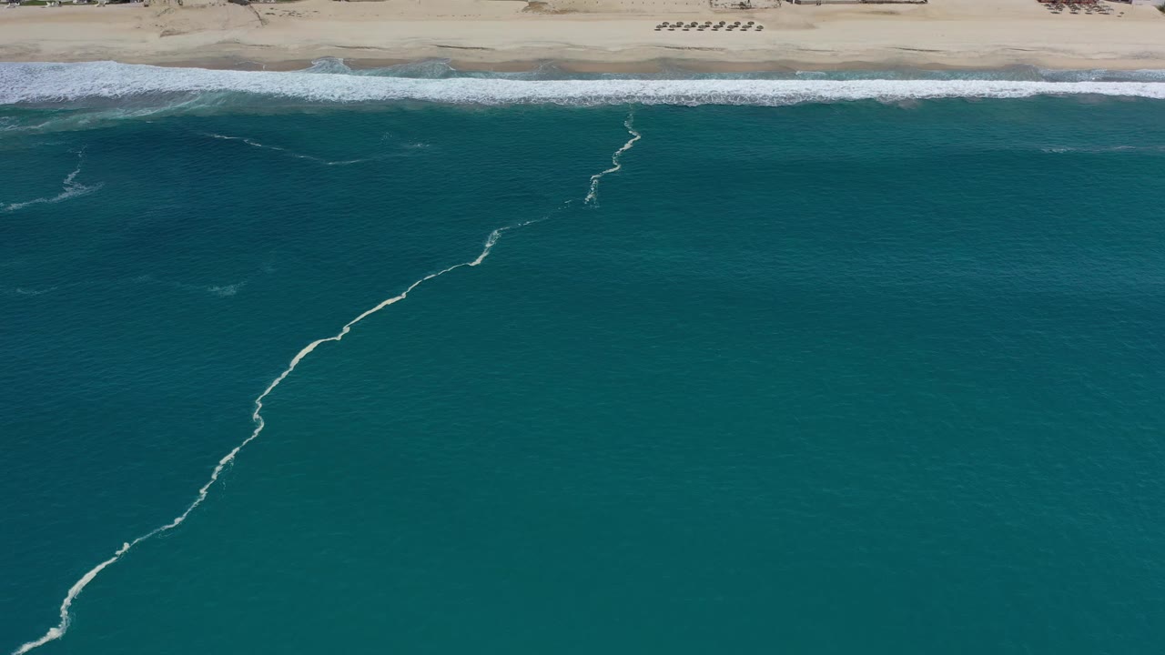 Aerial frame over Los Cabos, Mexico with blue-green water filling the view, the sandy beach held at the top, a broken white foam trail in the water, and no visible people.