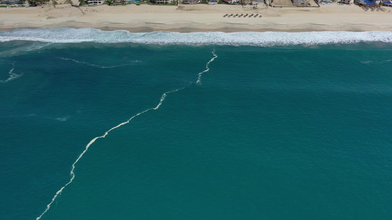 High aerial view of teal ocean and a sandy Los Cabos beach, with a thin white foam line angling through the water and no visible people.