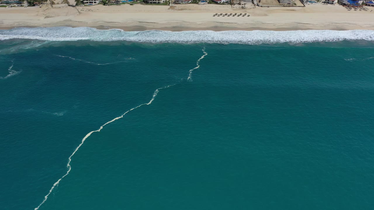 Aerial Los Cabos shoreline frame with deep teal water below, white surf across the beach, and a small row of beach umbrellas or loungers near the sand, with no visible people.