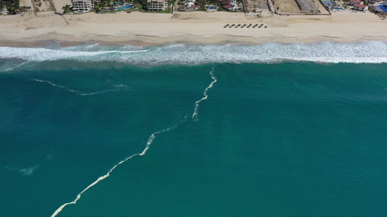 Aerial view over Los Cabos, Mexico with sea-green water, white surf lines, tan beach, small beachfront buildings, and no visible people.