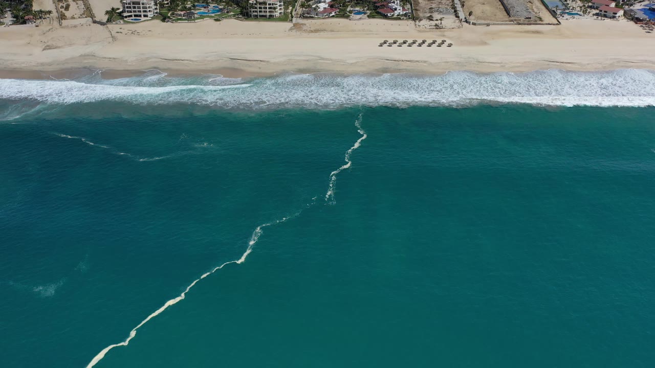 Overhead Los Cabos coastline frame with a diagonal foam streak in sea-green water, surf breaking near the sand, beach chairs above, and no visible people.