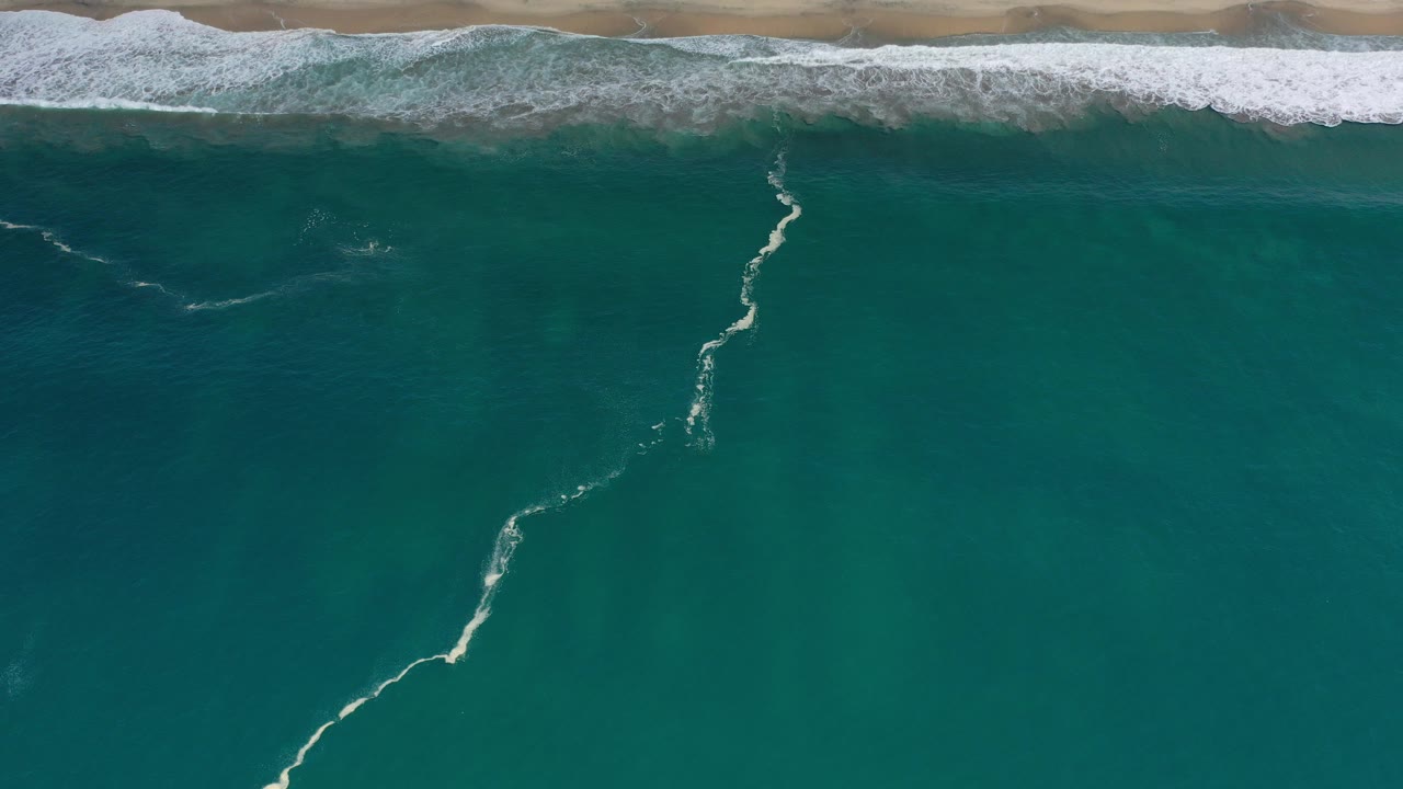 Overhead teal ocean with white surf near a sandy beach.