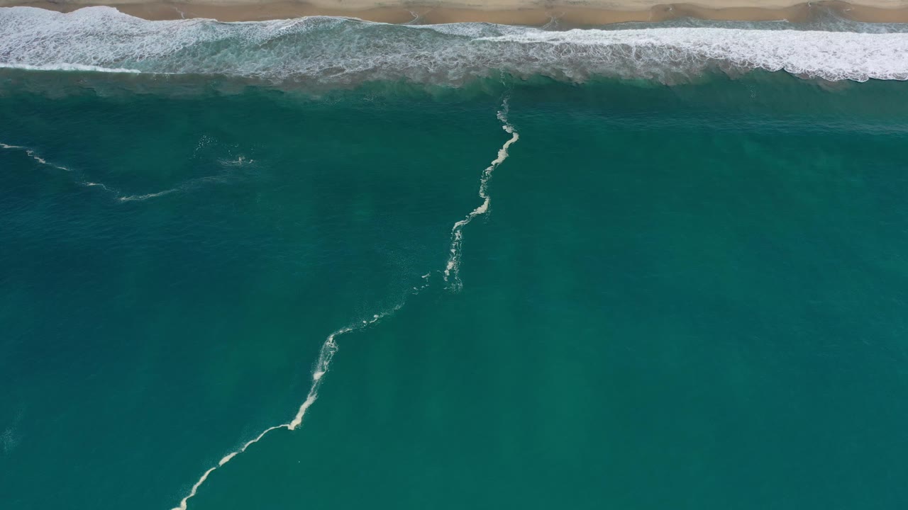 Top-down Los Cabos shoreline with a thin white foam trail running through green-blue water toward the breaking surf, with no visible people.