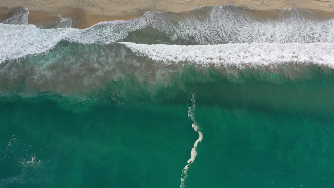 Overhead view in Los Cabos, Mexico, with teal ocean below, a white surf band across the center, tan sand at the top, and no visible people.