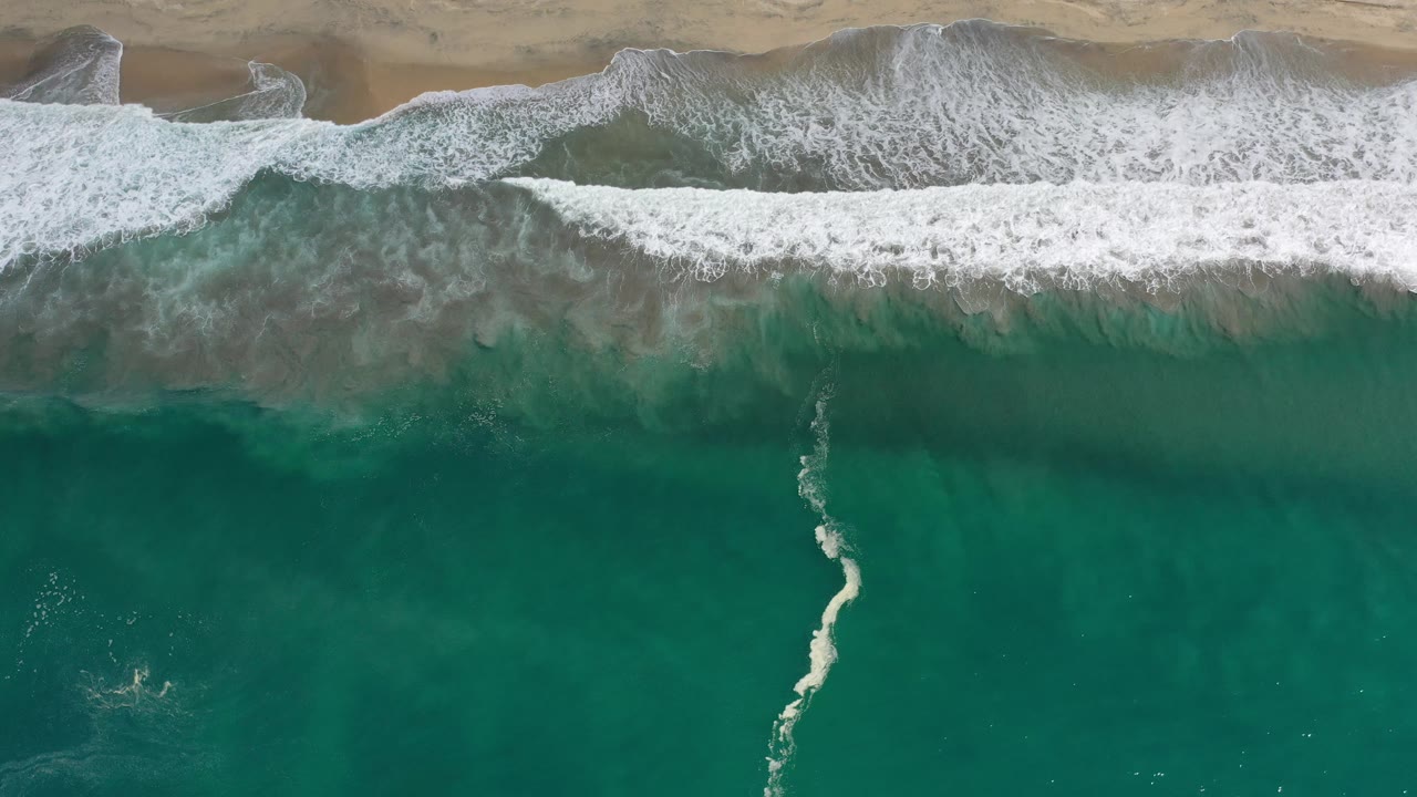Aerial Los Cabos beach frame with a wider sheet of white foam pushing toward the tan shoreline and darker green water in the lower half, with no visible people.