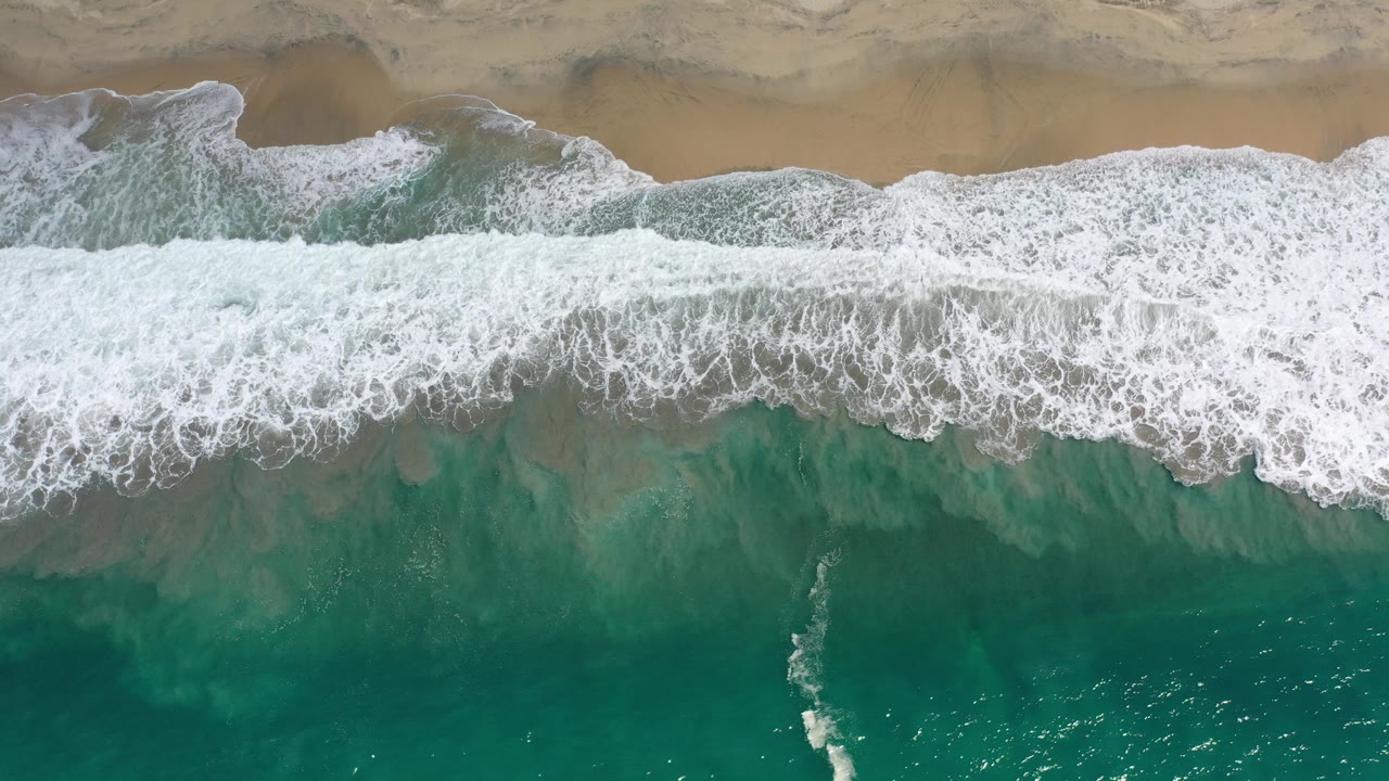 Top-down view of blue green water and white surf reaching a tan Los Cabos beach at the start of the clip, with no visible people.